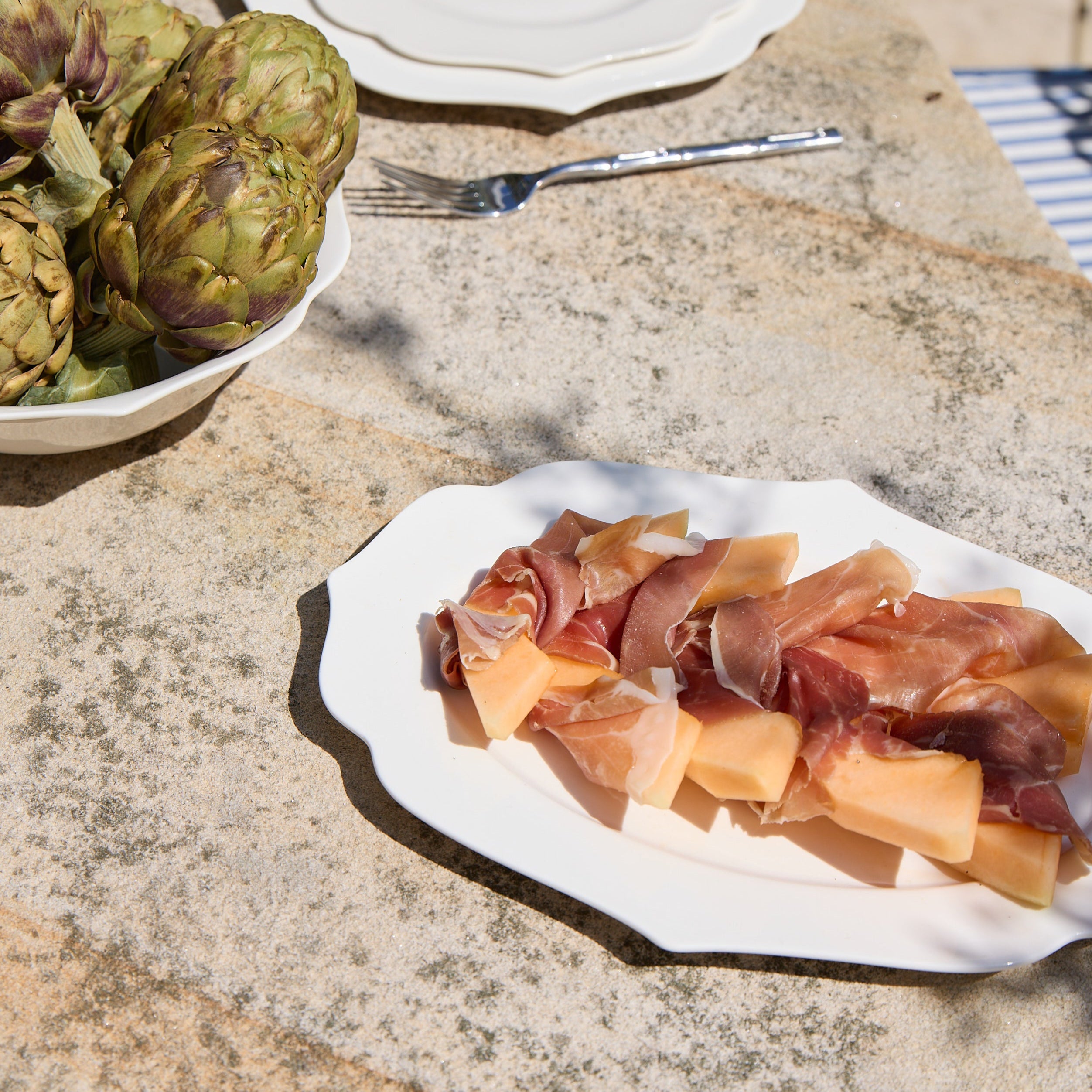 Platter of prosciutto and melon on a stone surface with artichokes and a plate in the background.