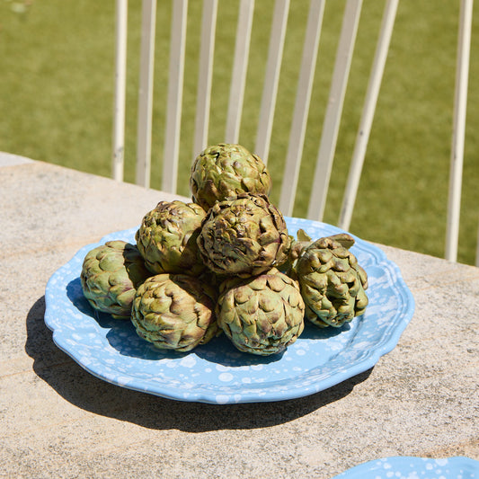 Blue plate with green artichokes on a stone surface with a white metal chair and grass in the background.