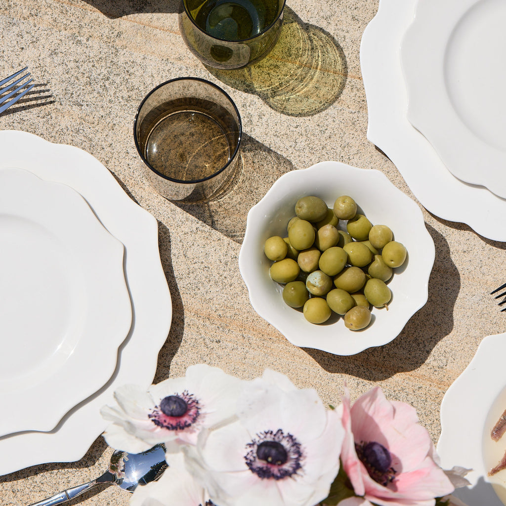 Dining table setting with white plates, silverware, and a bowl of olives on a textured surface.