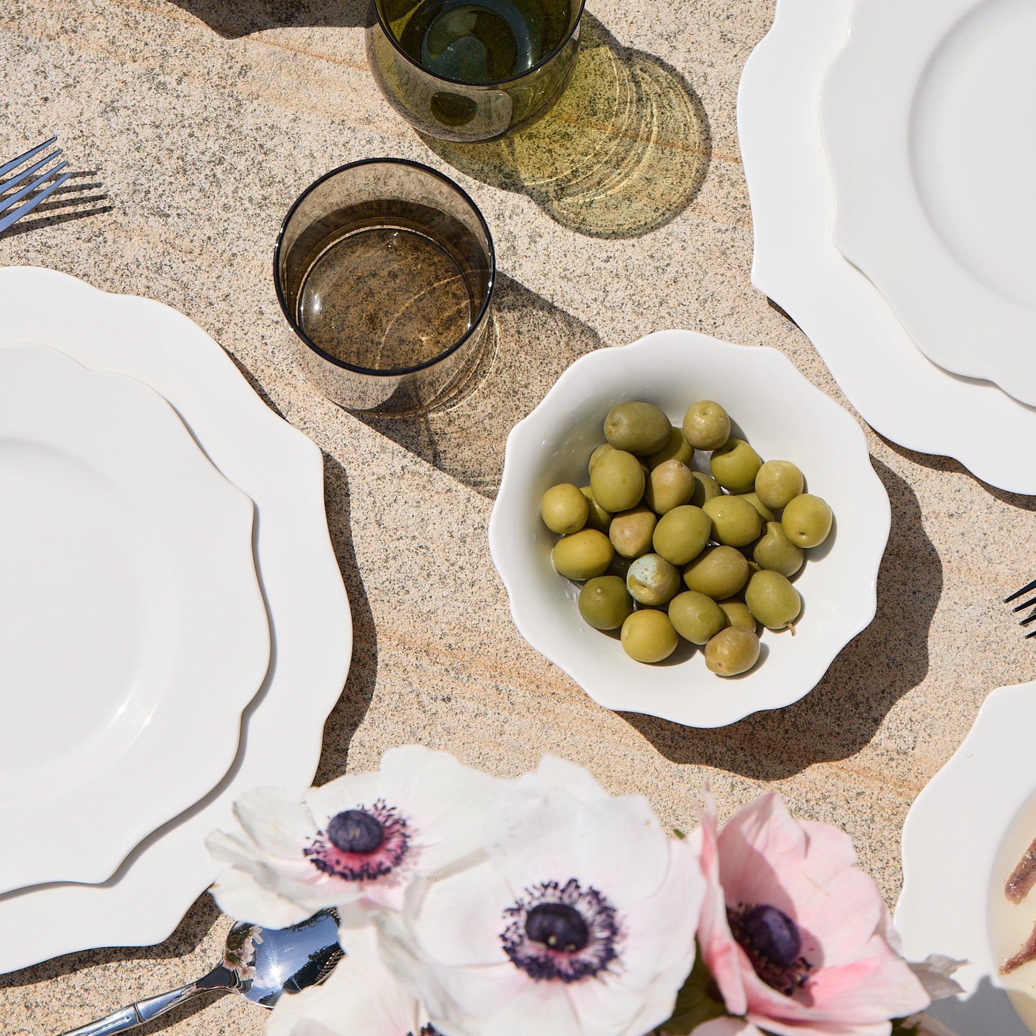 Dining table setting with white plates, silverware, and a bowl of olives on a textured surface.