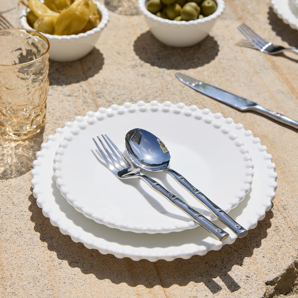 Set table with white plates, silverware, and small bowls of olives and pickles on a stone surface.