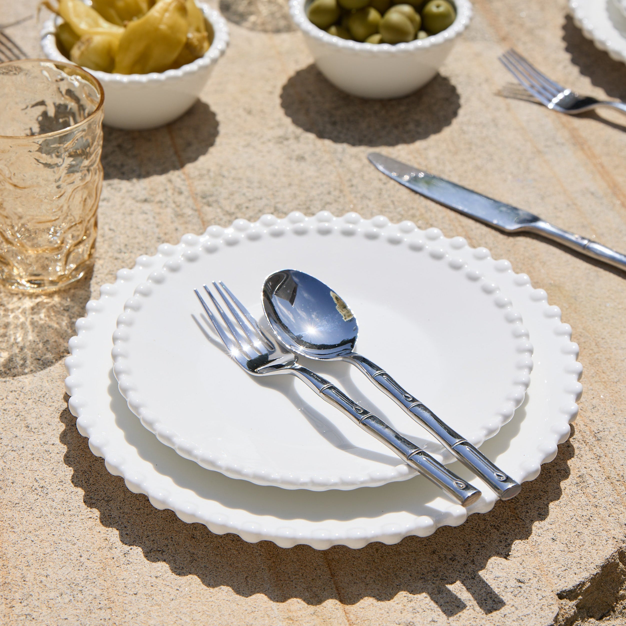 Set table with white plates, silverware, and small bowls of olives and pickles on a stone surface.