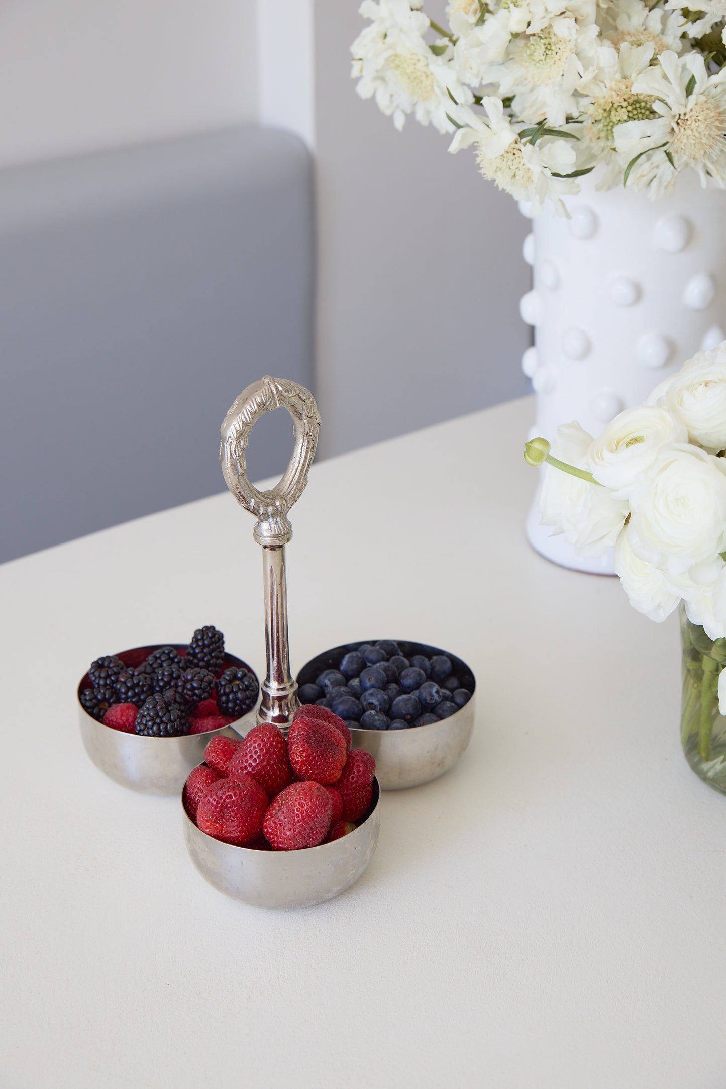 Silver berry holder with red and black berries on a white surface with flowers in the background