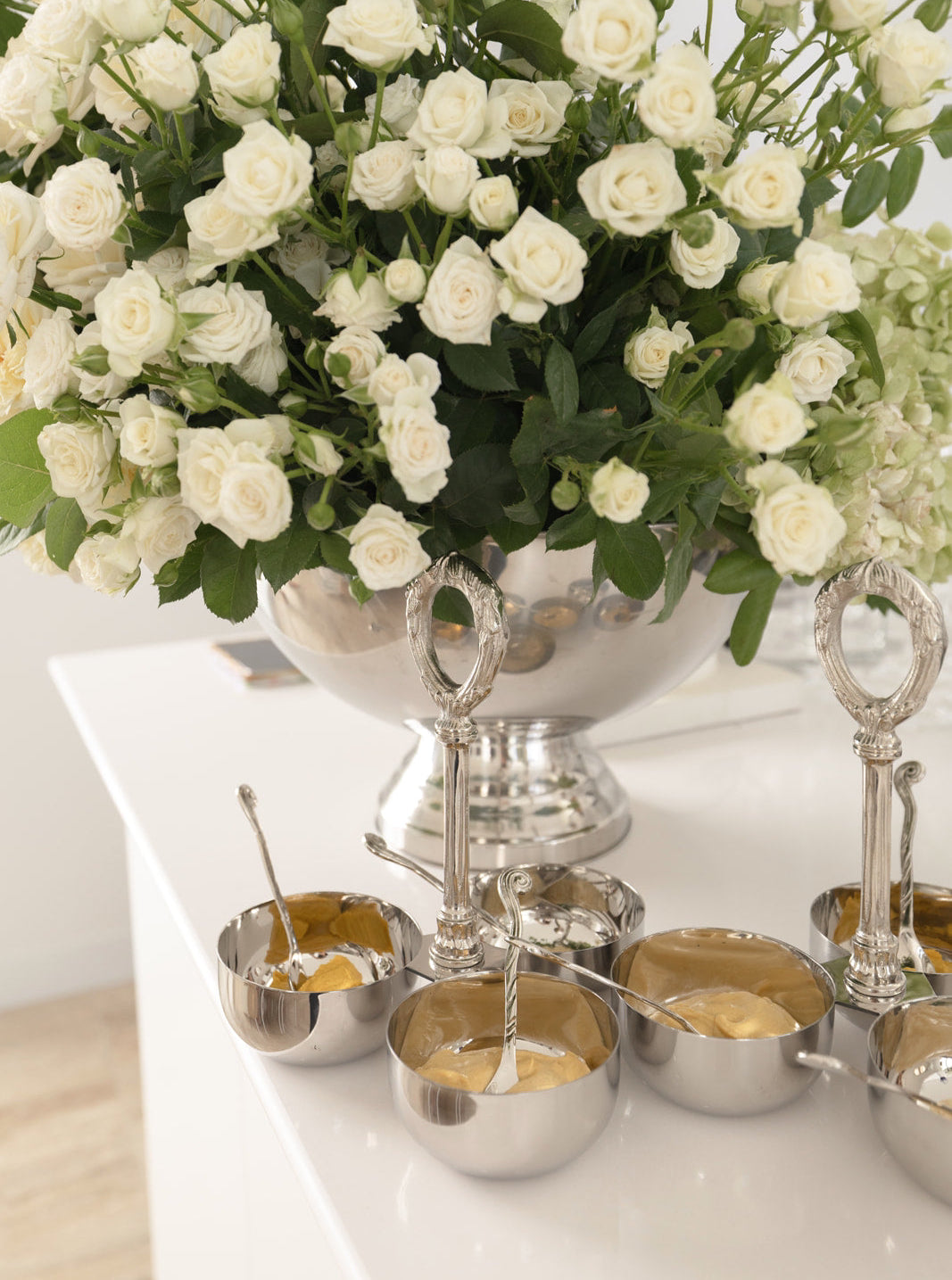 White flowers in a silver vase with silver bowls and spoons on a white surface