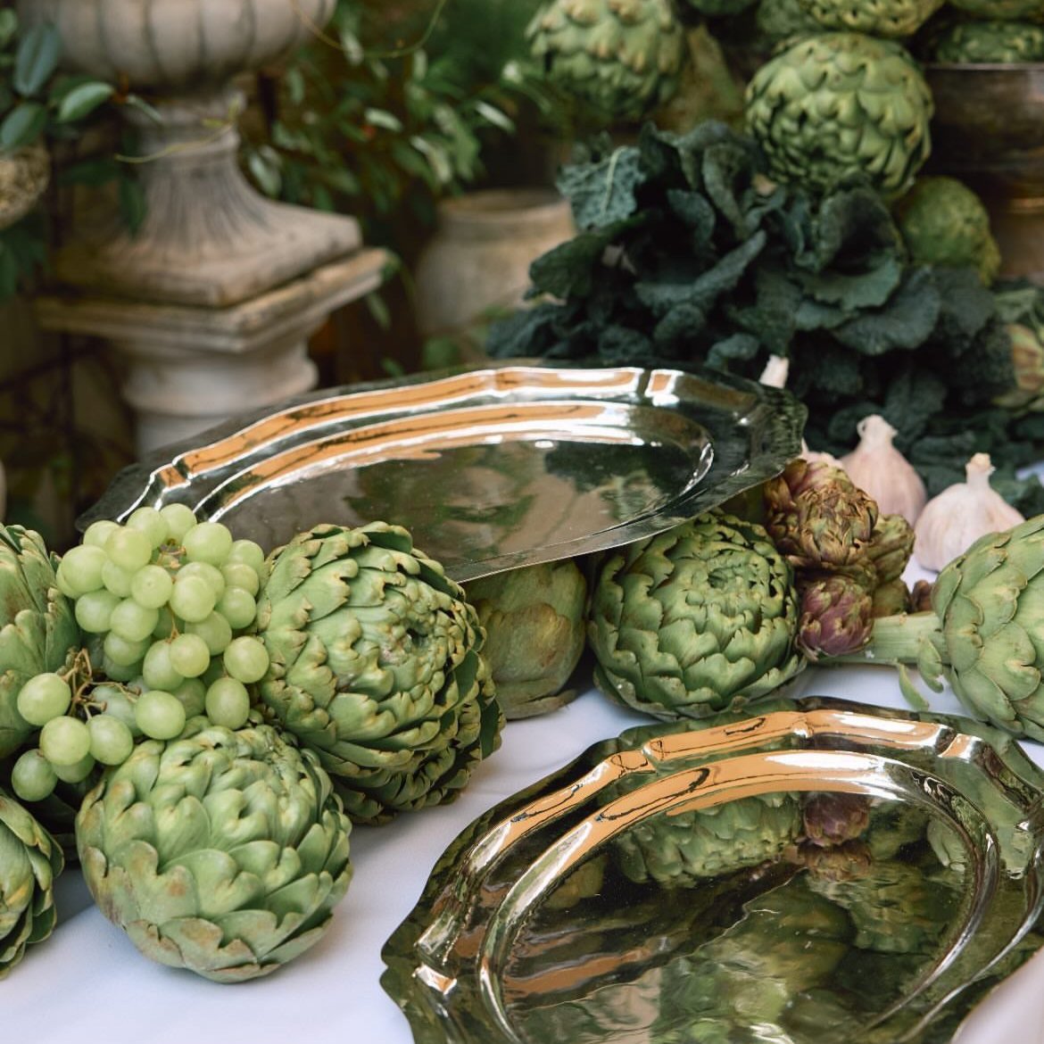 A silver serving tray with a reflective surface, placed among green vegetables and fruits, indicating the tray is used for serving.