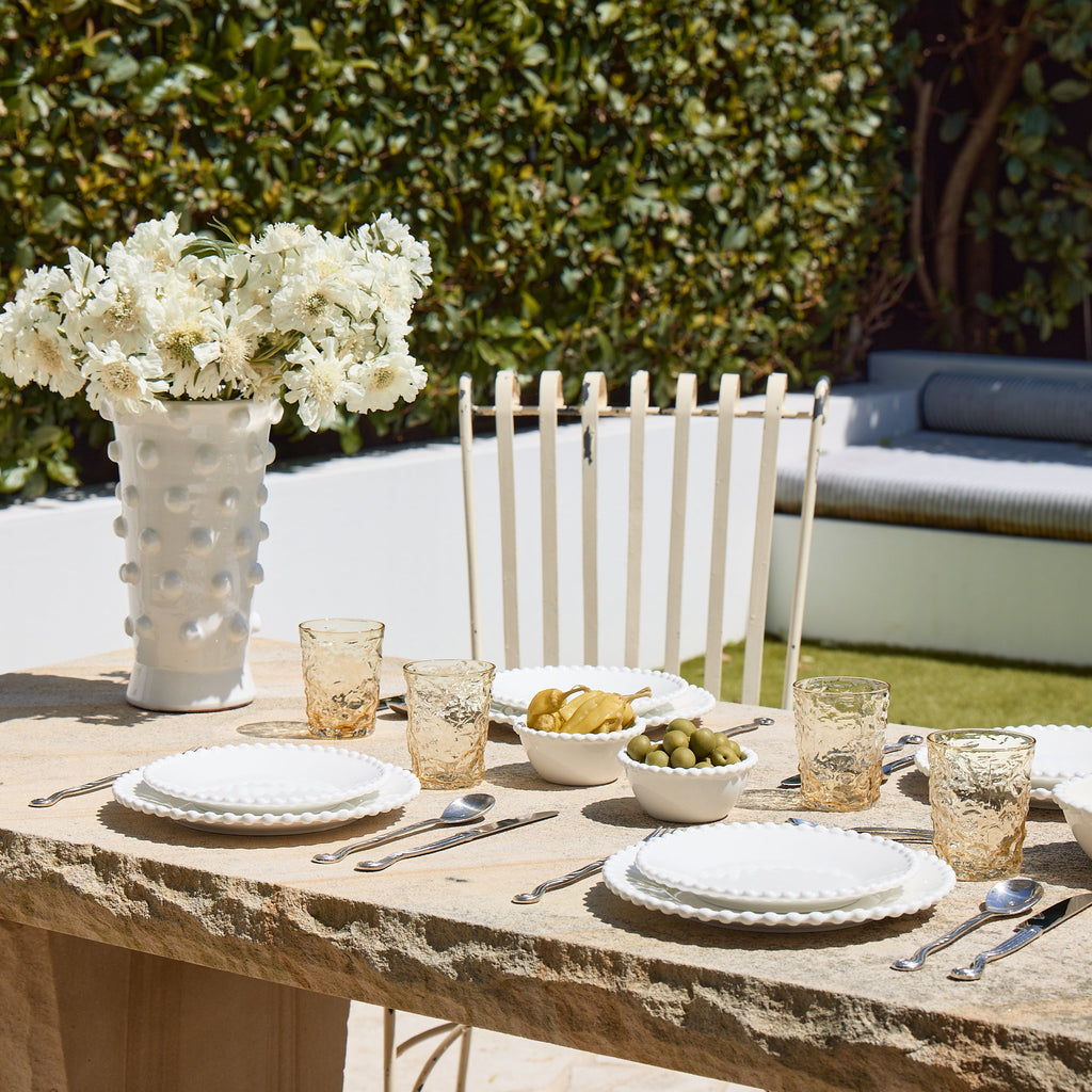 Outdoor dining setup with a stone table, white chairs, and a vase of white flowers.