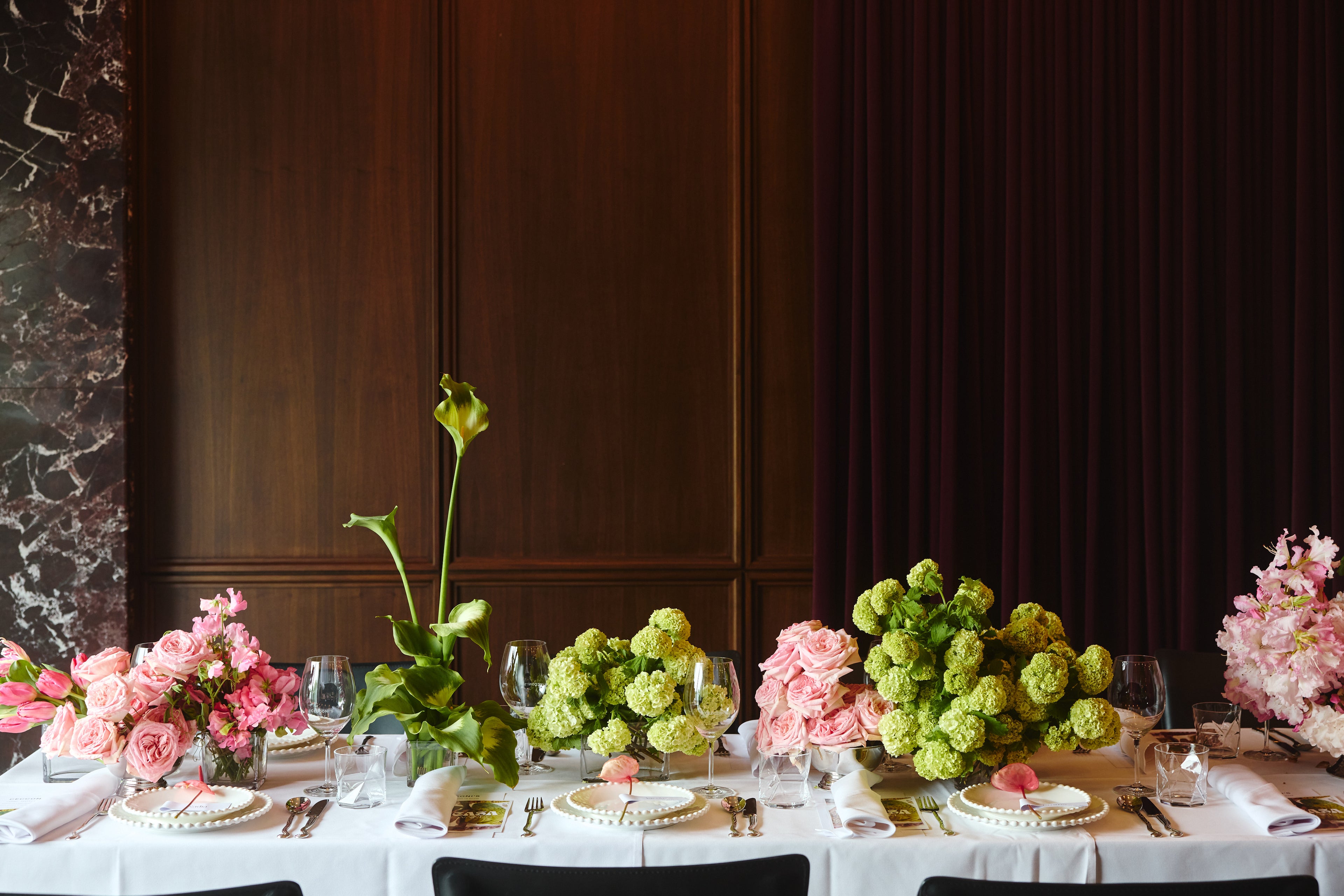 A long dining table set with white tablecloths, place settings, wine glasses, and floral centerpieces featuring pink flowers and green hydrangeas, set against a dark wood-paneled wall.
