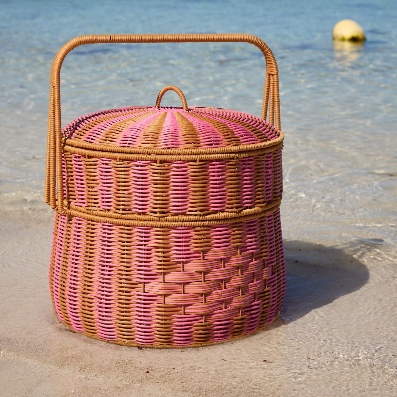 Wicker picnic basket with a pink lid on a sandy beach with clear blue water and green buoys in the background.